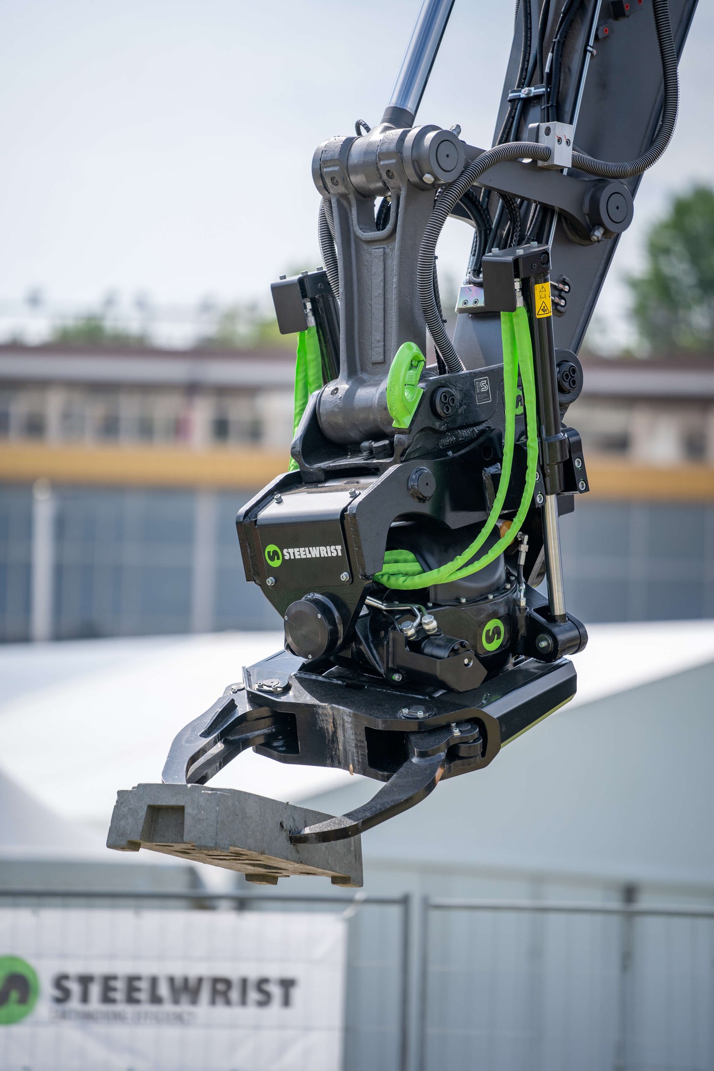 A close-up of a Steelwrist industrial excavator arm attachment with a steel grab claw, featuring black and gray metal components with bright green hydraulic hoses and branding.